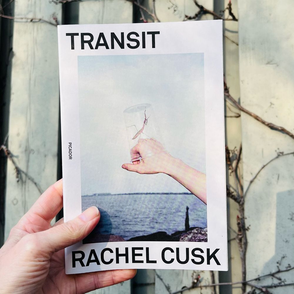 A white hand holds a copy of the book Transit by Rachel Cusk. A beige fence is in the background with leafless vines climbing up it. The book has a white cover with black text. In the middle of the cover is a photo of a white hand in front of some kind of water in the background (a lake maybe?). A praying mantis is sitting on top of the hand and it has been covered with a clear plastic disposable cup. 