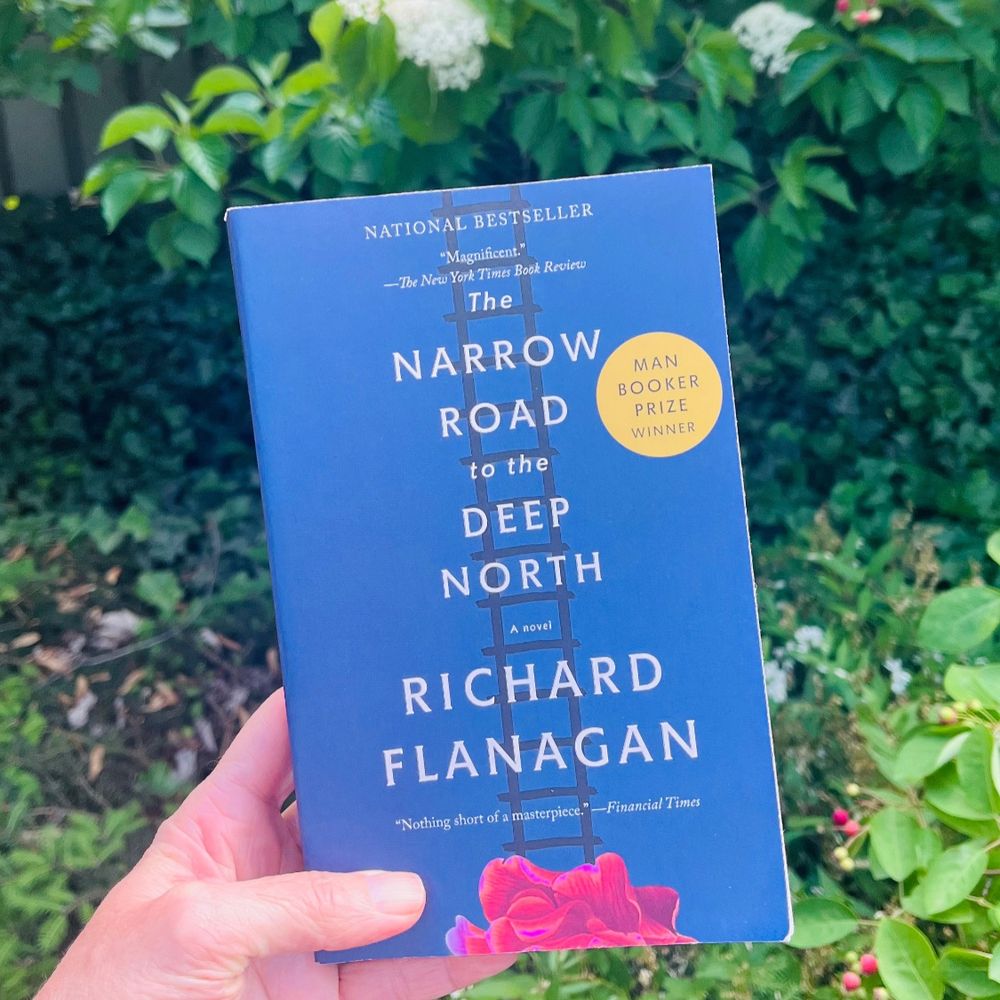 A white hand holds the book Narrow Road to the Deep North by Richard Flanagan in front of a lot of green shrubs with white flowers and red berries. The book is blue with a ladder, a pinkish red flower, and a yellow circle that says “Man Booker Prize Winner”. 