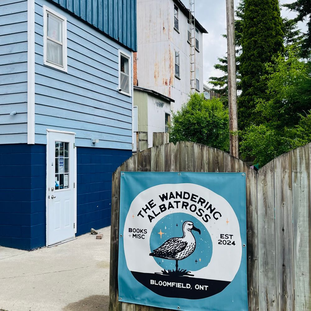 The outside of a building which is dark blue below and light blue above. In front is a fence that has a sign for The Wandering Albatross Books in Bloomfield, Ontario. There is a black and white drawing of an albatross (which looks like a super-sized gull) in the middle. 