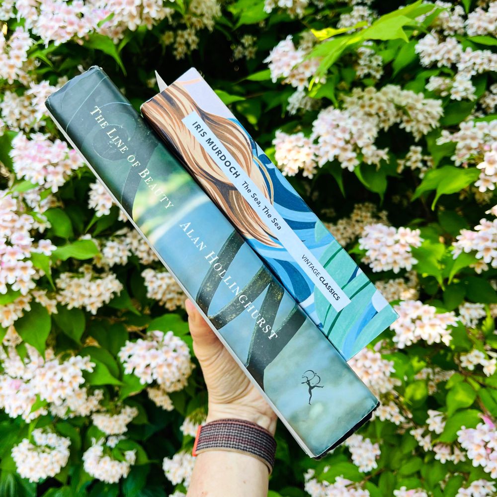 A white hand holds two books in front of a flowering shrub. The books are Alan Hollinghurst’s The Line of Beauty and Iris Murdoch’s The Sea, The Sea. Both books have blueish-greenish covers. 