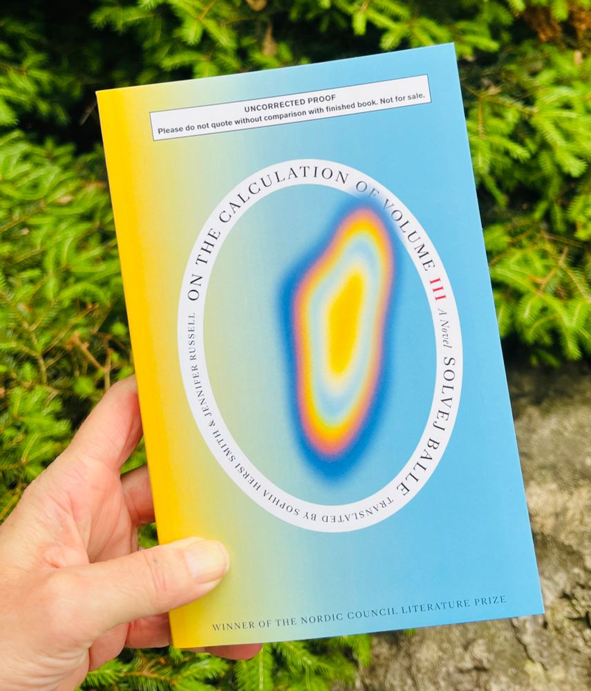 A white hand holds a blue and yellow book above a rock and some green shrubs. The book is an uncorrected proof of On the Calculation of Volume Book 3 by Solvej Balle and translated by Sophia Hersi Smith. The book cover has a multicolored blob in the middle and this is encircled by a white oval which contains the text. 