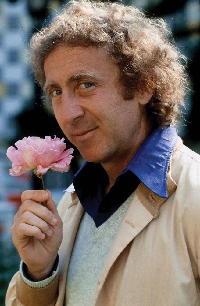 Gene Wilder, looking adorably sidelong at the camera, holding a pink flower wearing a tan jacket, taupe vest, and cornflower blue collared shirt