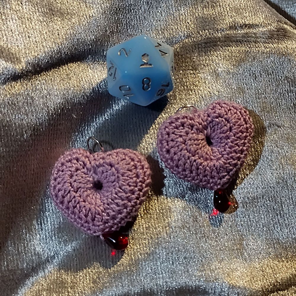 Two crochet heart pendants in light purple with a single red teardrop glass bead at the bottom of each. With d20 for scale.