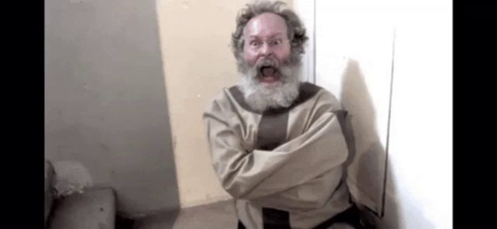 a man with a beard and a straitjacket is sitting in a room with his arms crossed .