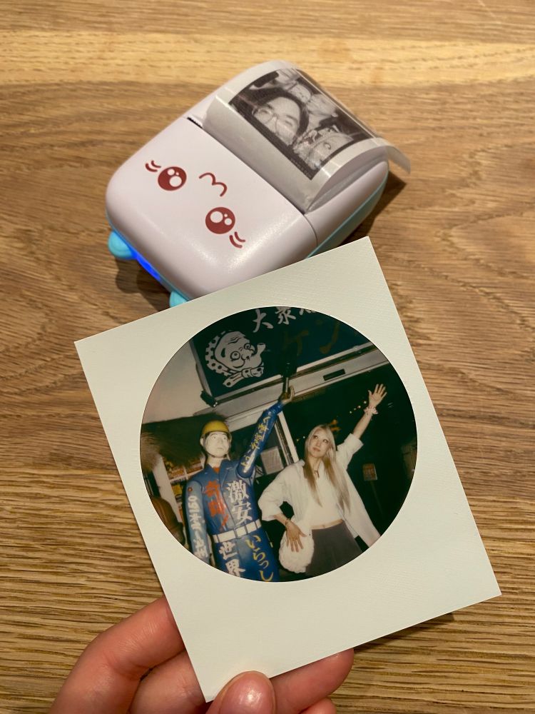 A round frame Polaroid of my friend Semi holding her left arm to the air, mimicking a statue. It lies next to her sticker printer, shaped like a cat with teal ears.