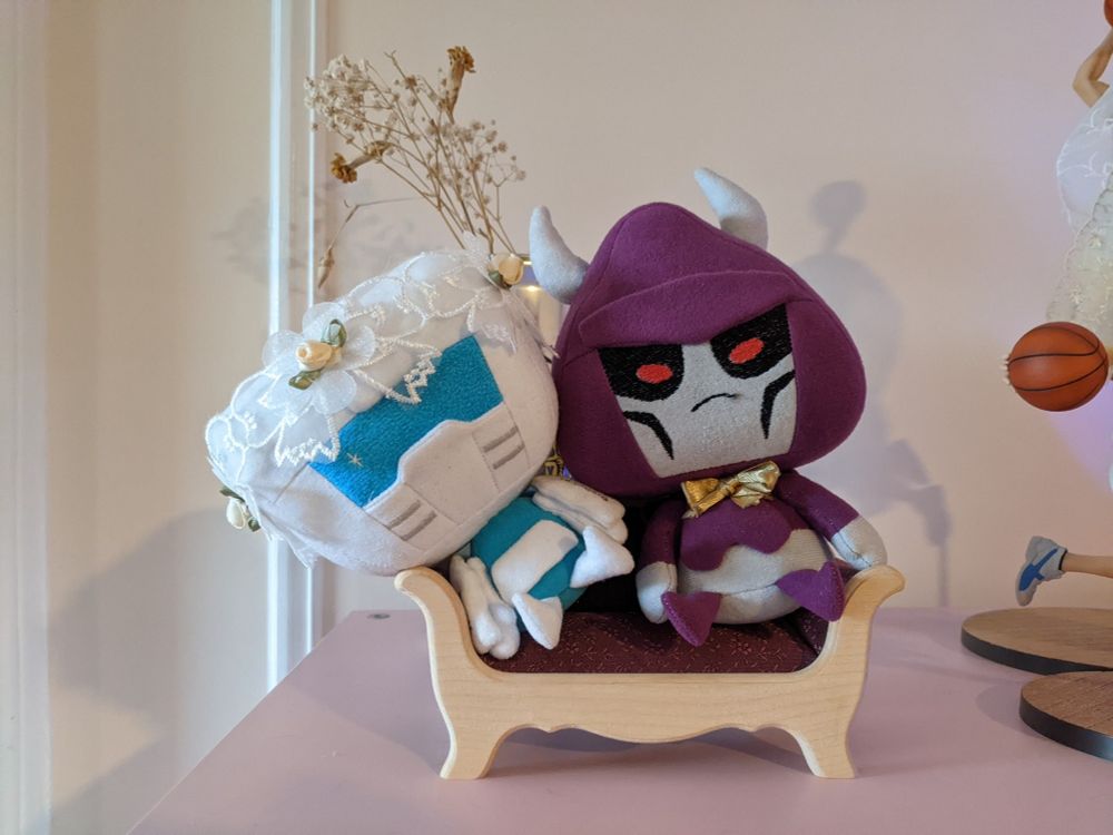 Tailgate and Cyclonus plush dolls sitting on a tiny couch. Tailgate has a bridal headpiece and Cyclonus has a bowtie
