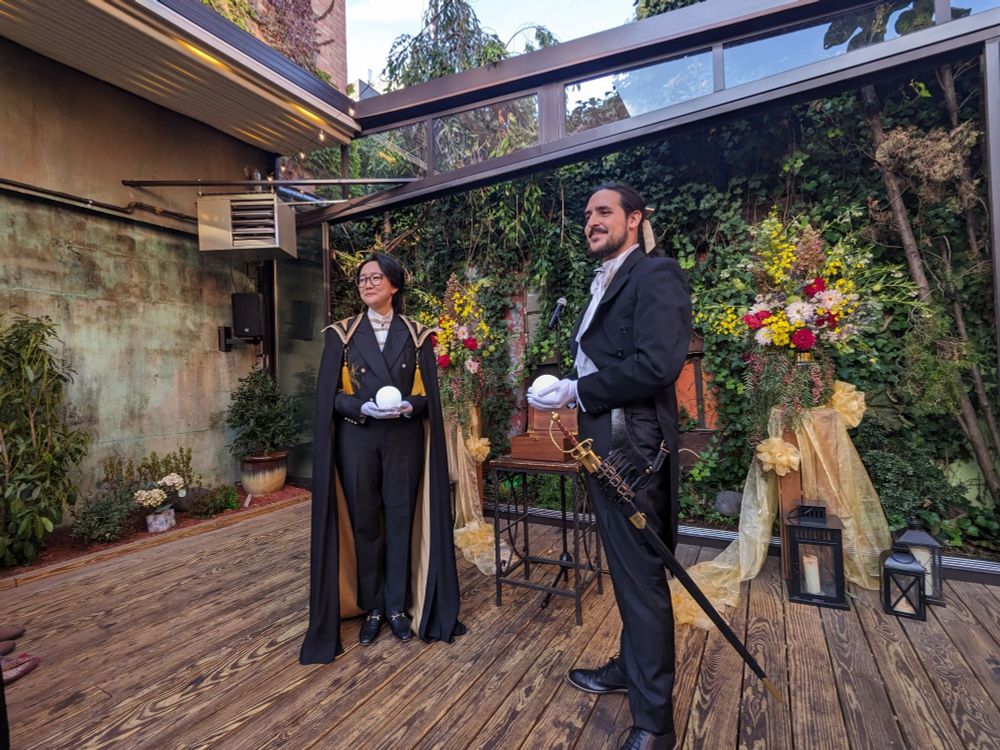 Photo of the grooms holding glowing "stars". Two men in suits, one in a cape and the other with a sword on his side, pose in front of two flower arrangements on a wooden outdoor deck at a Spanish-themed restaurant.