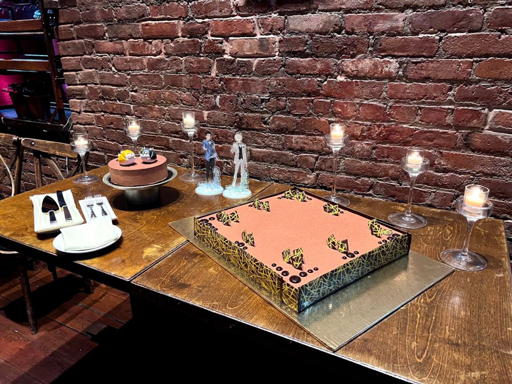 Two cakes on display. The round cake has toppers of two round Shiba Inu (yellow and brown), and acrylic standees of Yuri and Victor stand between the cake.