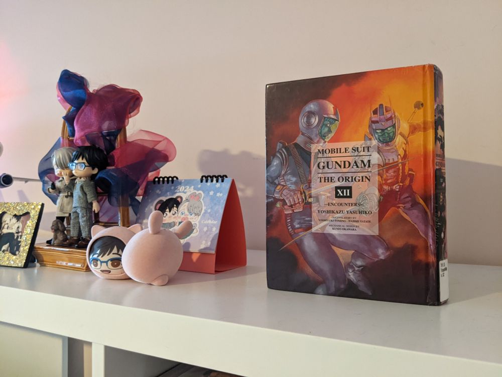 Bookshelf display of Gundam The Origin Pt. XII (featuring Amuro and Char) and assorted Yuri On Ice decorations