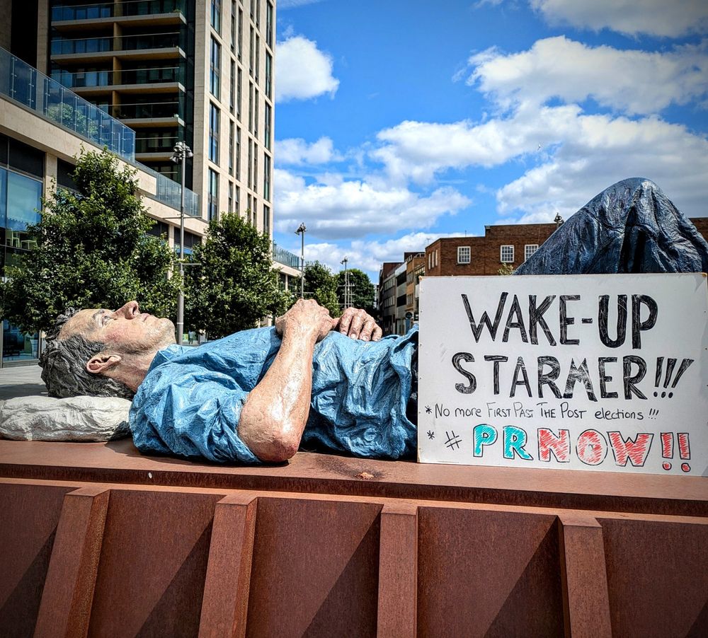 Medium shot photo of Artist Sean Henry's 'Catafalque (lying man)' on a iron oxide plinth outside the tower blocks in central Woking.  A sculpture of a giant man asleep with his knees raised.  MVMNORTHWESTSURREY have placed a placard with the following phrase at the figure's legs: "WAKE-UP STARMER!!!  NO MORE FIRST PAST THE POST ELECTIONS.  PR NOW!!" Part of the local group's contribution to the Democracy Action weekend - marking one year since the most disproportional election in UK history.