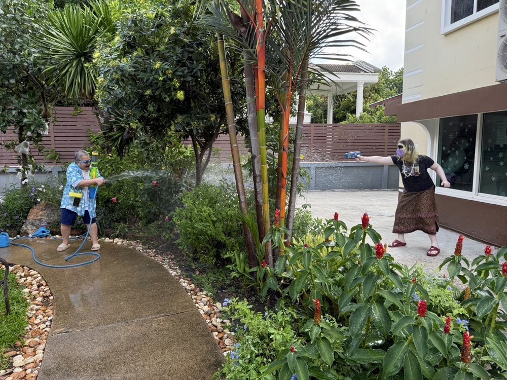A photo of two people in a garden, having a water fight. The person on the right is holding a water gun, wearing sunglasses, a face mask, a necklace.