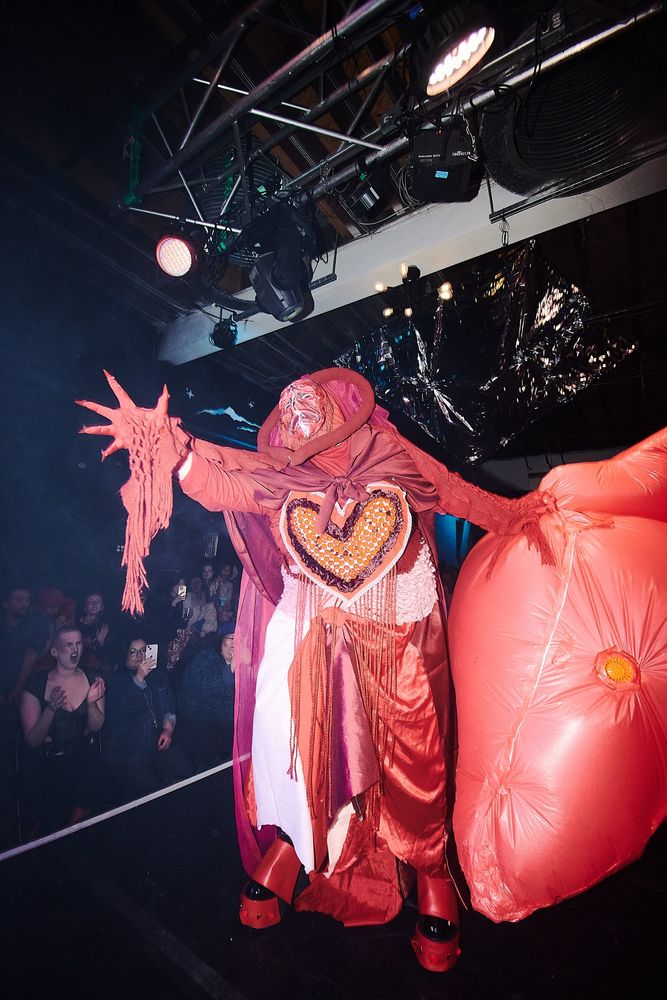 A costumed figure in various shades of red fabric standing on a stage. There is a heart shape on their chest and they are holding a giant red inflatable heart. One gloved hand is outstretched towards the top left corner