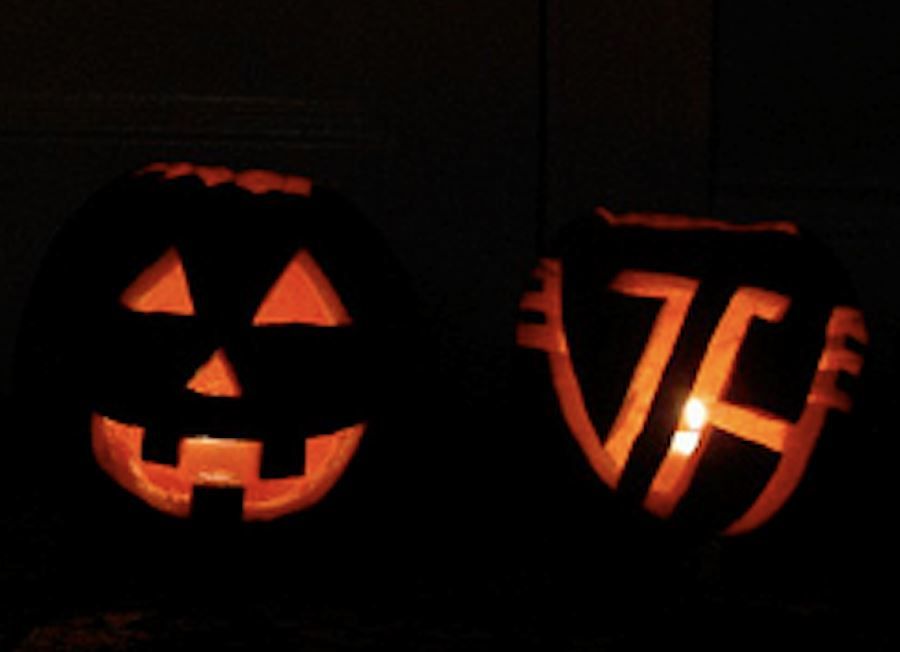 Close-up of two carved pumpkins lit from the inside by candles in a dark room. The carving on the left is a traditional Halloween pumpkin face design. The carving on the right is the Van Halen logo.