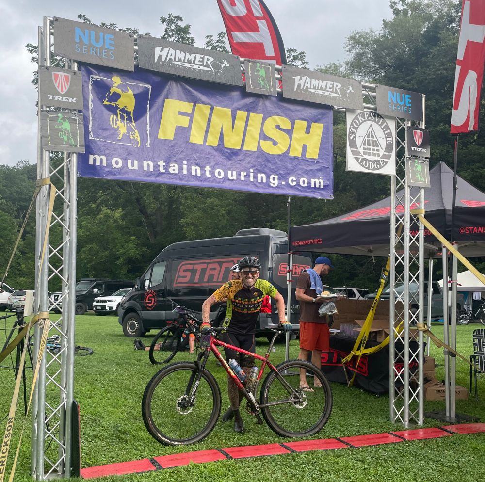 A very muddy man standing with a red mountain bike at the finish line of a mountain bike race facing the camera and smiling. 