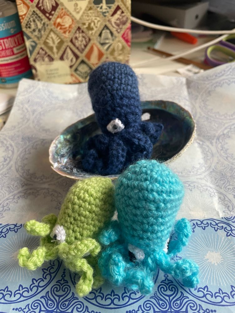 A green, a pale blue and a navy crocheted octopuses sitting in a Paua shell 