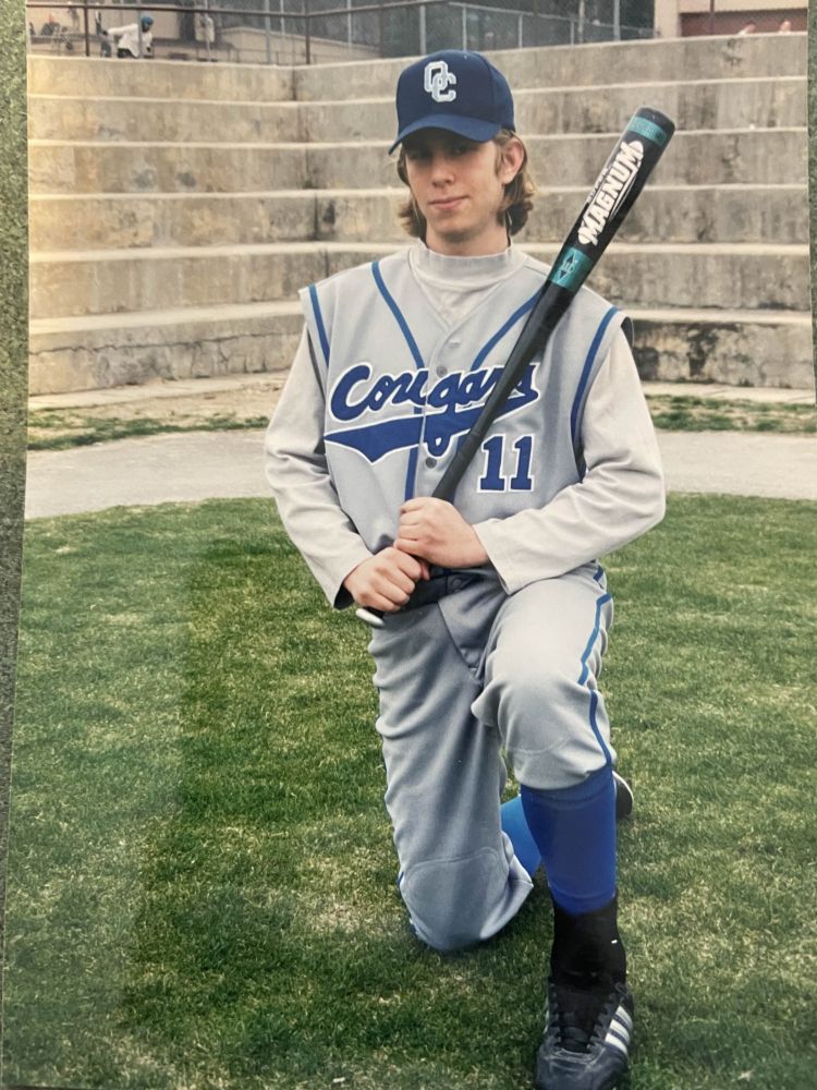 Portrait of a high school baseball player kneeling with a bat over his left shoulder. His long, golden hair complements his fierce determination to one day rediscover this photo while cleaning out his garage. 