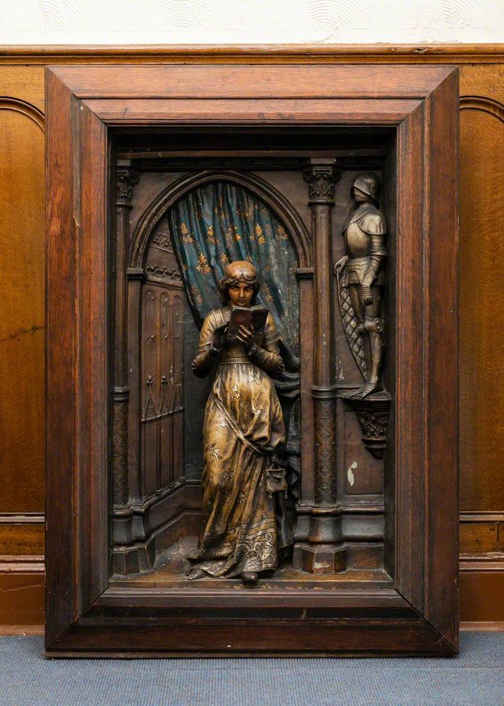 A hand-painted bronze plaque in a wooden frame. It shows a young woman standing in a long dress in a doorwa with a suit of armour to the right and and blue and gold cloth behind her. She is reading a book which she holds in both hands.