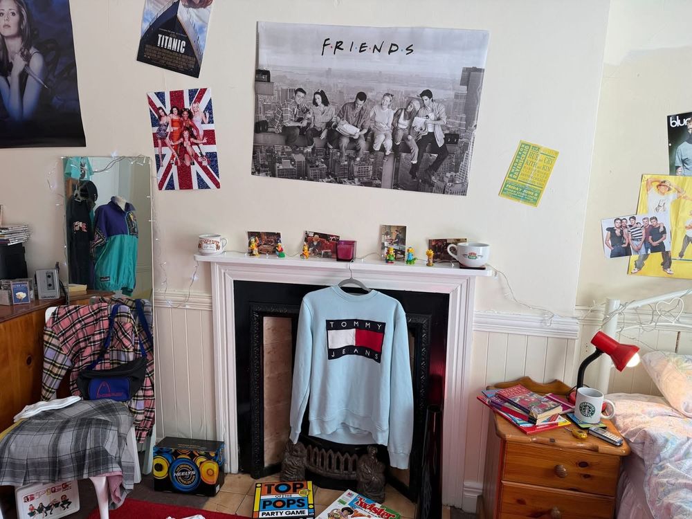 Part of a mocked-up 1990s young person's bedroom with a 'Friends' poster over the firelace and clothing hanging up.