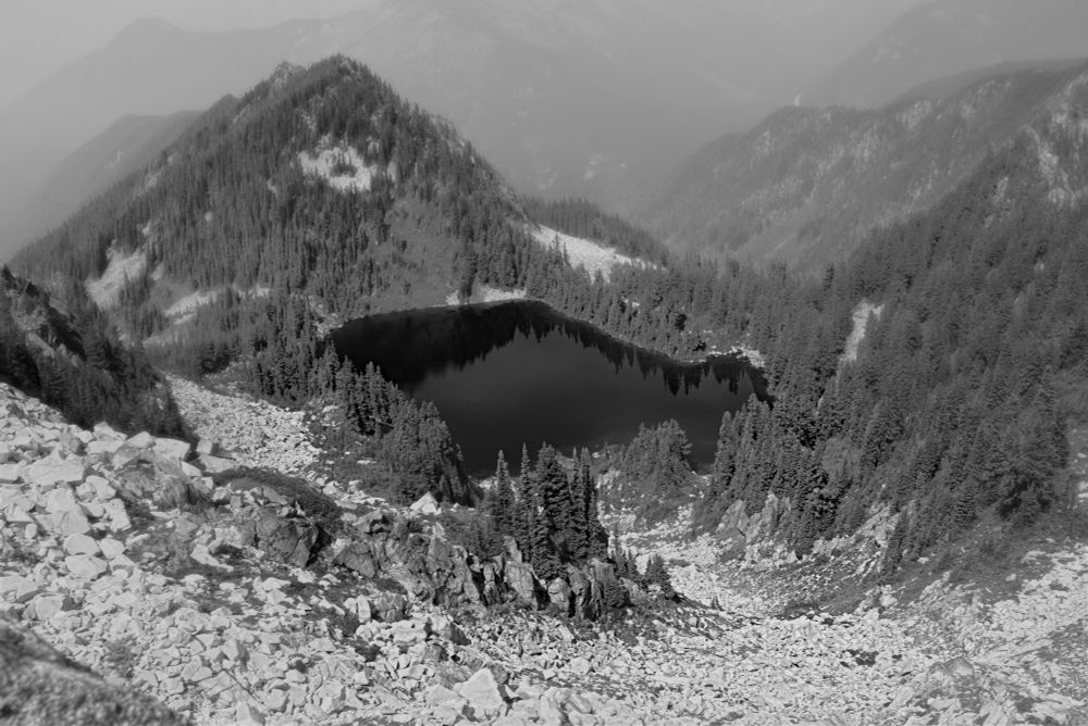 A mountain lake rimmed with conifers sits in a bowl in the Cascade Mountains.  A scree slope leads steeply downward to the lake while wildfire smoke obscures the mountains behind the lake