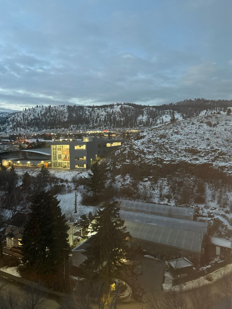 Picture of TRU. Some buildings in the foreground. Snow covered hills in the background. Just before dawn