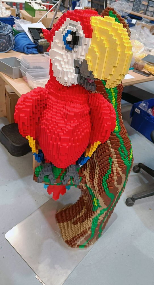 A cartoony scarlet macaw made entirely out of LEGO bricks. It's mainly red with a big yellow beak and white face. Sitting on a tree branch.