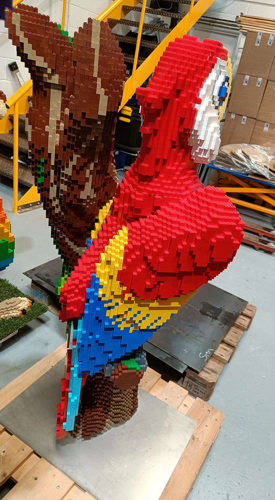 A cartoony scarlet macaw made entirely out of LEGO bricks. It's mainly red with a big yellow beak and white face. Sitting on a tree branch.
A side view showing the blue and yellow on the wings.