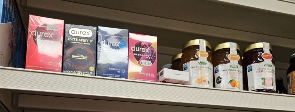 A photo of a shop shelf with a selection of condoms sitting alongside jars of no added sugar jam