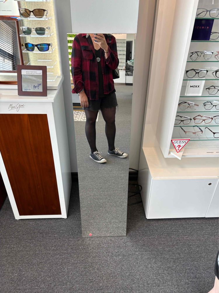 Angela in a full-length mirror, with her head cropped off. She’s wearing an open plaid button-up over a black tunic top that has moon on it. Black cut-off shorts, black pantyhose, and black and white Chucks. She’s in an optometrist’s office with glasses frames on shelves 