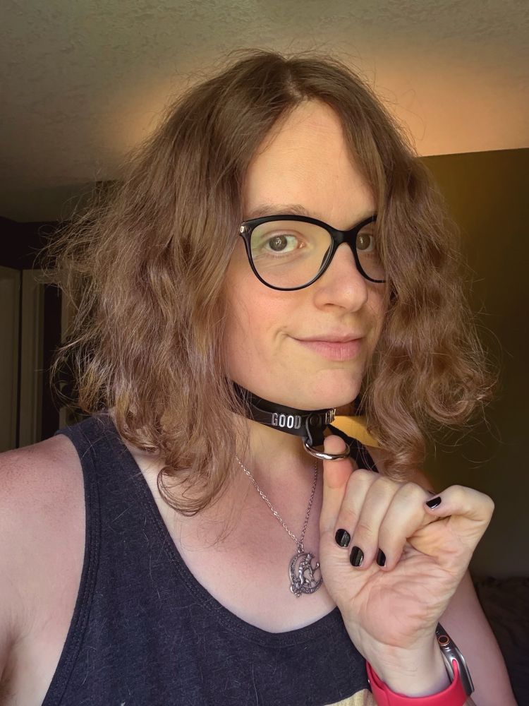 Angela tugging happily at the ring on a black leather collar that says “GOOD GIRL” in silver letters 