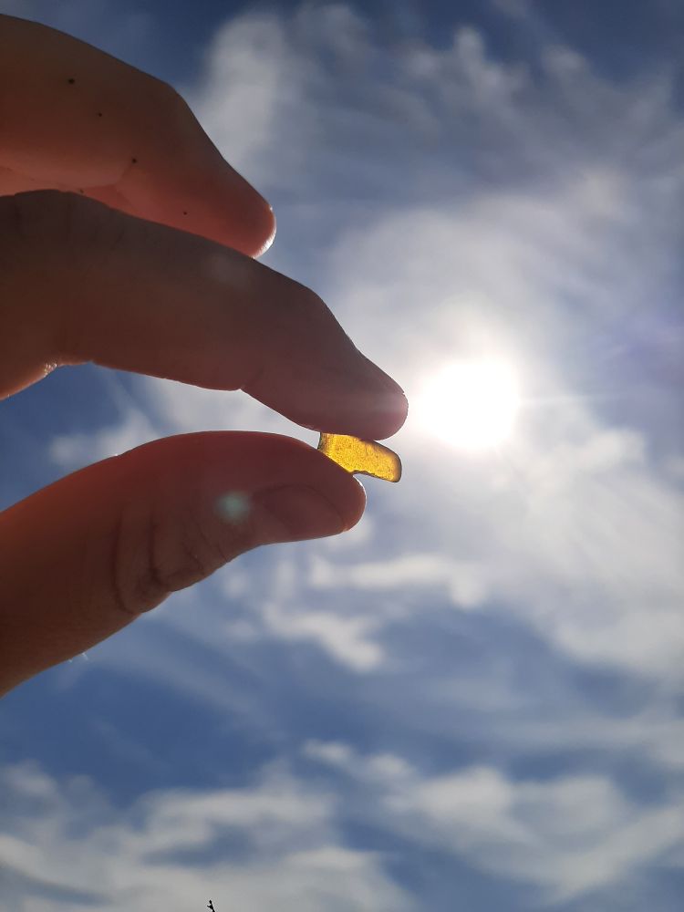 A rectangular piece of brown sea glass being held up to the sun. It glows gold with the sun's light.
