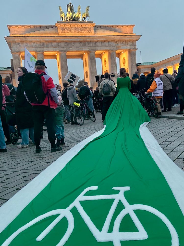Eine Frau, die von hinten zu sehen ist, trägt ein grünes Kleid mit engem, ihren Körper betonenden Oberteil mit einer langen Schleppe. Die Schleppe stellt einen Radweg dar, zu erkennen an den „Verkehrszeichen“: weißen „Seitenstreifen“ und einem großen Fahrrad-Piktogram. 