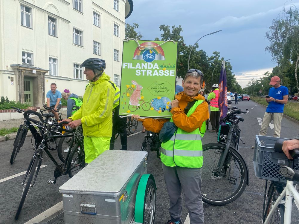 Auf der Fahrbahn der Filandastraße hält eine Fahrrad-Aktivistin bei der Zwischenkundgebung einer Demo für sichere Radwege auf der Filandastraße in Berlin Steglitz-Zehlendorf ein buntes Plakat mit dem Straßenamen hoch 