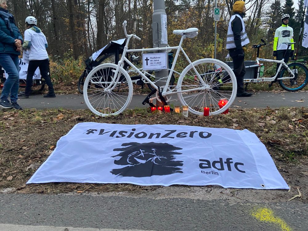 Geisterrad mit Kerzen und VisionZero Fahne des ADFC Berlin am Unfallort Müggelheimer Damm Ecke Müggelhort, in der Nähe des Müggelsees. Die Straße ist von einem Wald umgeben, es ist herbstliches Laub zu sehen und einige Teilnehmende der VisionZero Demo am 09.11.2025