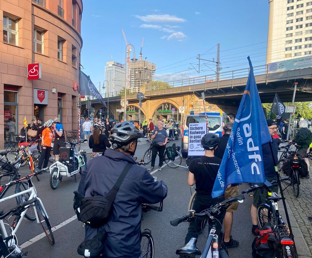 Auf der Fahrbahn am Hackeschen Markt in Mitte steht eine Gruppe Radfahrende bei einer Zwischenkundgebung der Respect Cyclists Demo. Im Vordergrund ist eine ADFC Berlin Fahne zu sehen, weiter im Hintergrund der weiße Aufsteller der Respect Cyclists mit schwarzer Schriftfarbe. 