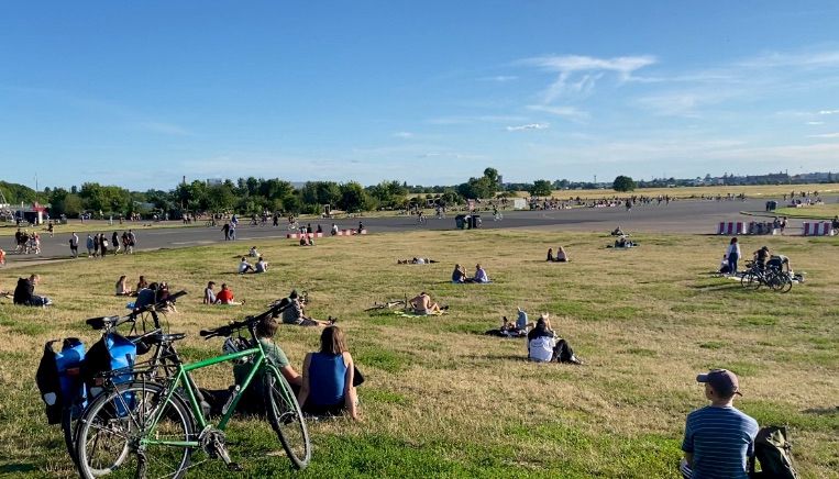 Tempelhofer Feld: Blick auf die Wiese vom Eingang Oderstraße Richtung Tempelhof. Der Horizont zeigt blauen Himmel ohne Häuser. Auf der Wiese sitzen Menschen in Grüppchen oder einzeln. Im Vordergrund steht ein grünes Fahrrad. 