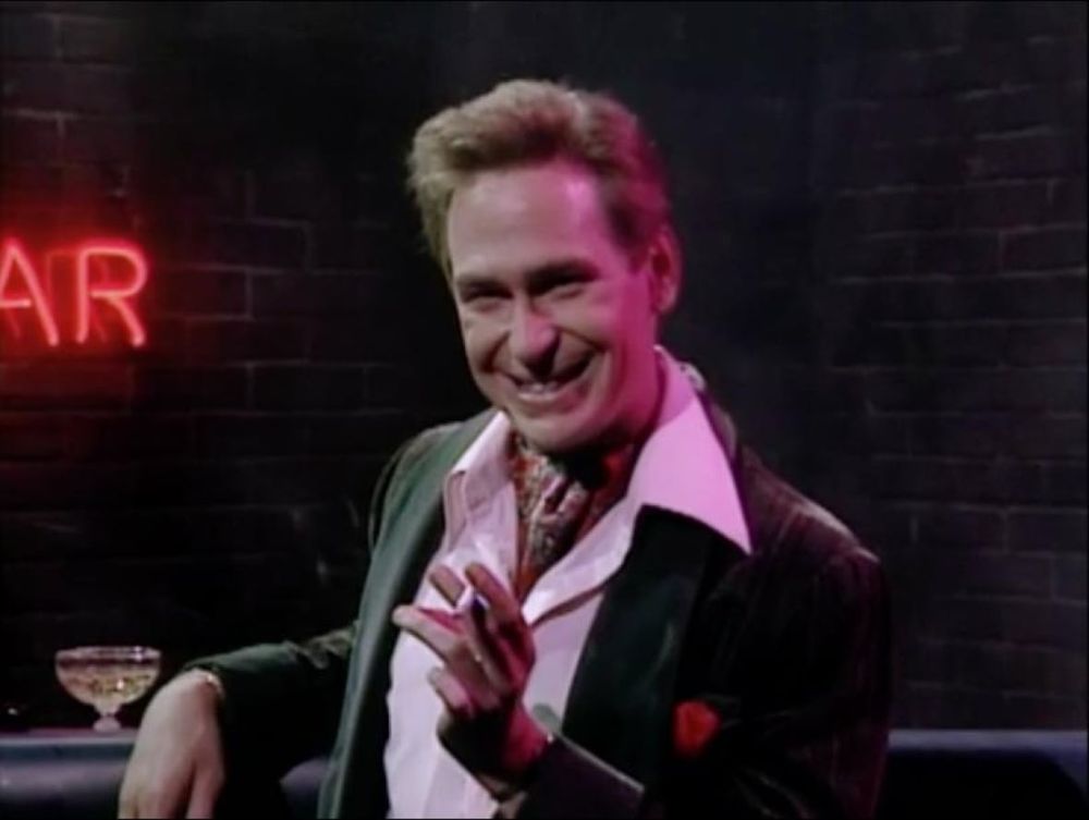 Scott Thompson as Buddy Cole, he is sitting at a bar with a cigarette in his left hand and he is grinning with great gay mischief. 