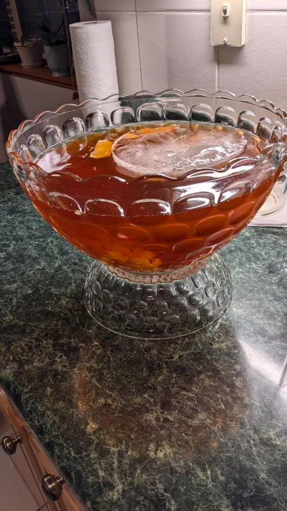 A glass bowl filled with a reddish brown liquid. A large, circular ice cube floats at the top along with a slice of orange peel.