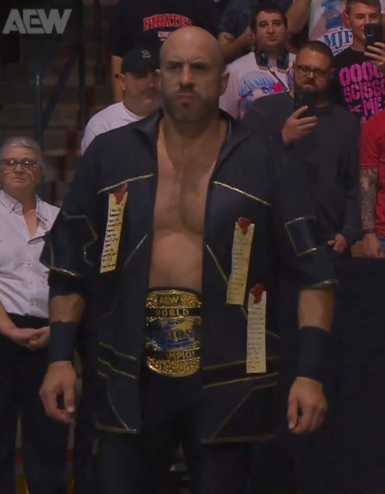 Professional wrestler Claudio Castagnoli, a massive slab of Swiss muscle. He wears a black and gold jacket over his Trios Championship belt. On his jacket are three Purity Seals--text on old-looking parchment strips affixed with red wax--from Warhammer 40k because he is a fucking nerd and that is why pro-wrestling is so excellent.