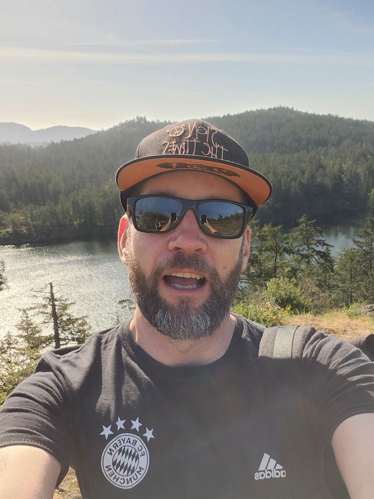The selfie of a dingus in a black shirt and sunglasses with a lake and trees in the background.
