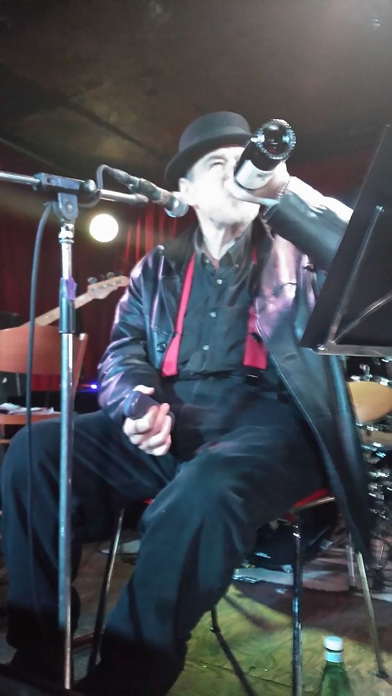A seated man at a microphone drinks from a wine bottle.