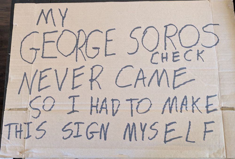 A brown cardborard sign with black marker reading as follows:
"My George Soros check never came so I had to make this sign myself."