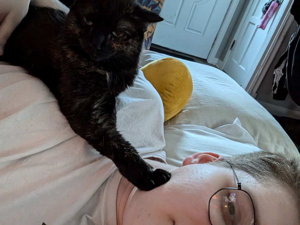 an older pic of me and mocha, with her laying on my chest, with half my face on the bottom right and mocha's face on the top left. in the middle, mocha's front paw reaches out and presses on my cheek. mocha's ears are laying flat and she is looking away from me.