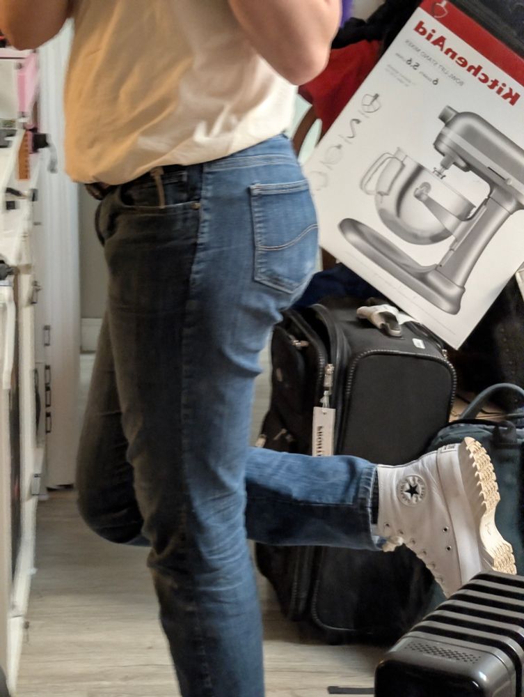 a mirror shot from my stomach down, showing my outfit with my leg lifted to show off my white converse. im wearing a white shirt and blue jeans. i am standing sideways so my butt's outline is visible. it is messy in the background. a suitcase and kitchenaid stand mixer box are prominent