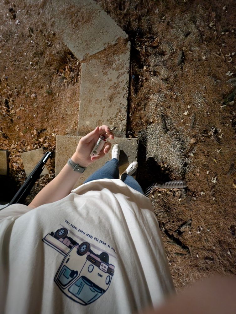 a top down shot from my upper chest showing my outfit. im standing outside on a small concrete pad by a sidewalk, wearing a white shirt and white shoes and blue jeans. in my visible hand im holding a joint and a white Plantasia lighter. the lighting is darker than usual and only from nearby street lamps