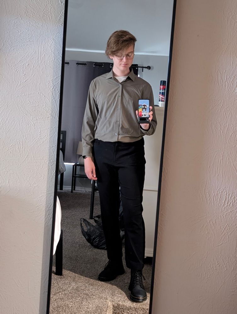 me standing in front of a full body mirror, taking a picture while holding my phone upside down and looking down at it. i'm wearing black doc marten's and black slacks, with an olive green button-down shirt tucked in