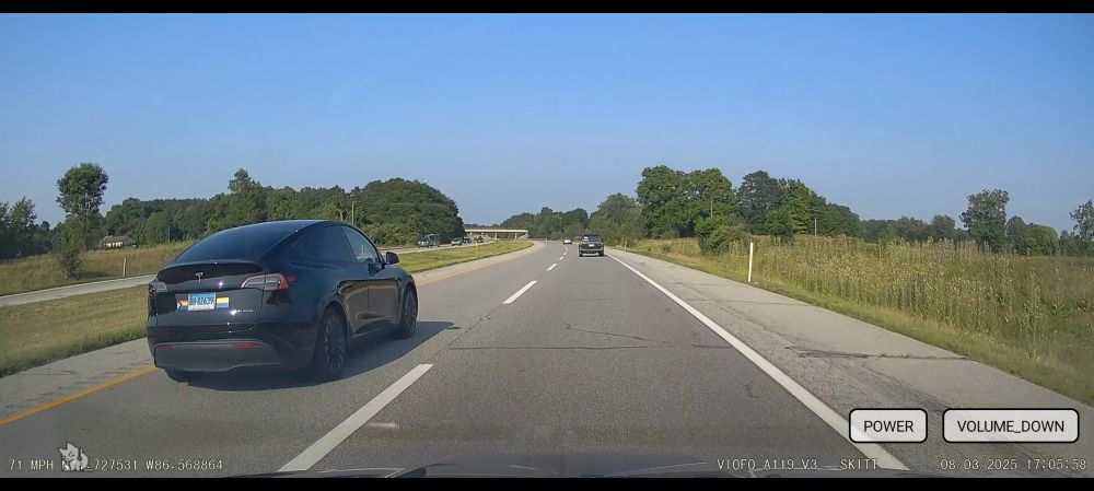 the view from my car's dashcam on the highway showing a black tesla in the left lane. it has a pride sticker or magnet on the back.