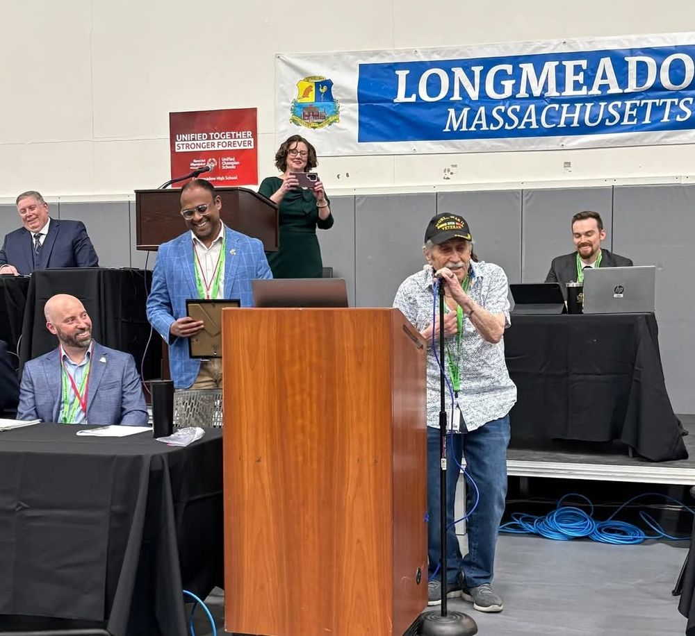 Man in a Veterans hat at a microphone behind a lectern speaks while 4 men and 1 woman look on smiling. The woman is taking a photo. 