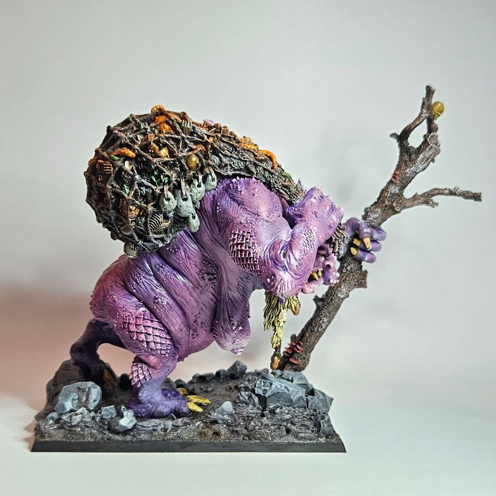 A warhammer old world Troll Hag in a walking pose facing right side on. Her skin is purple and pink, with dirty blonde hair. She is walking with a tree branch as a walking stick in her left hand and a fishing net/sack in her right tossed over her shoulder. She is very nude, this view gives a fairly obvious few of her rather large mammelrys.