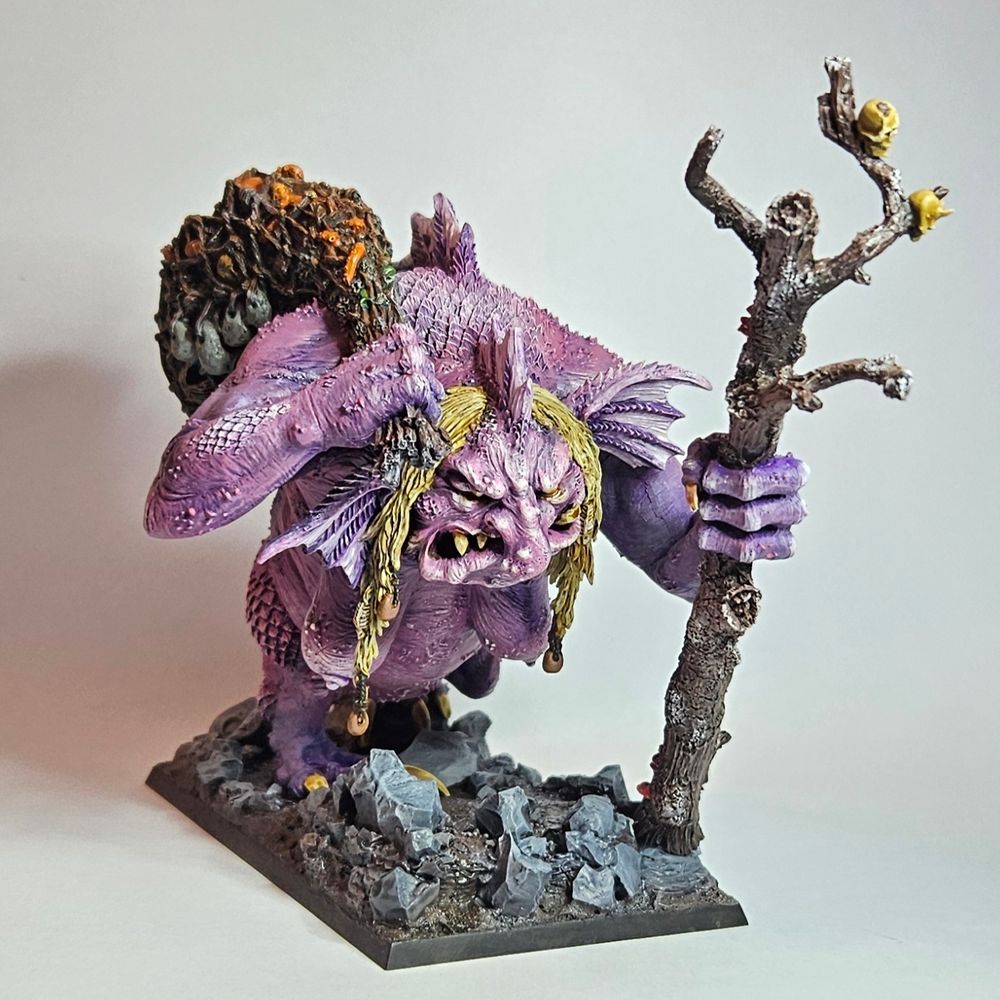 A warhammer old world Troll Hag in a walking pose facing front 3/4 on. Her skin is purple and pink, with dirty blonde hair. She is walking with a tree branch as a walking stick in her left hand and a fishing net/sack in her right tossed over her shoulder. She is very nude.