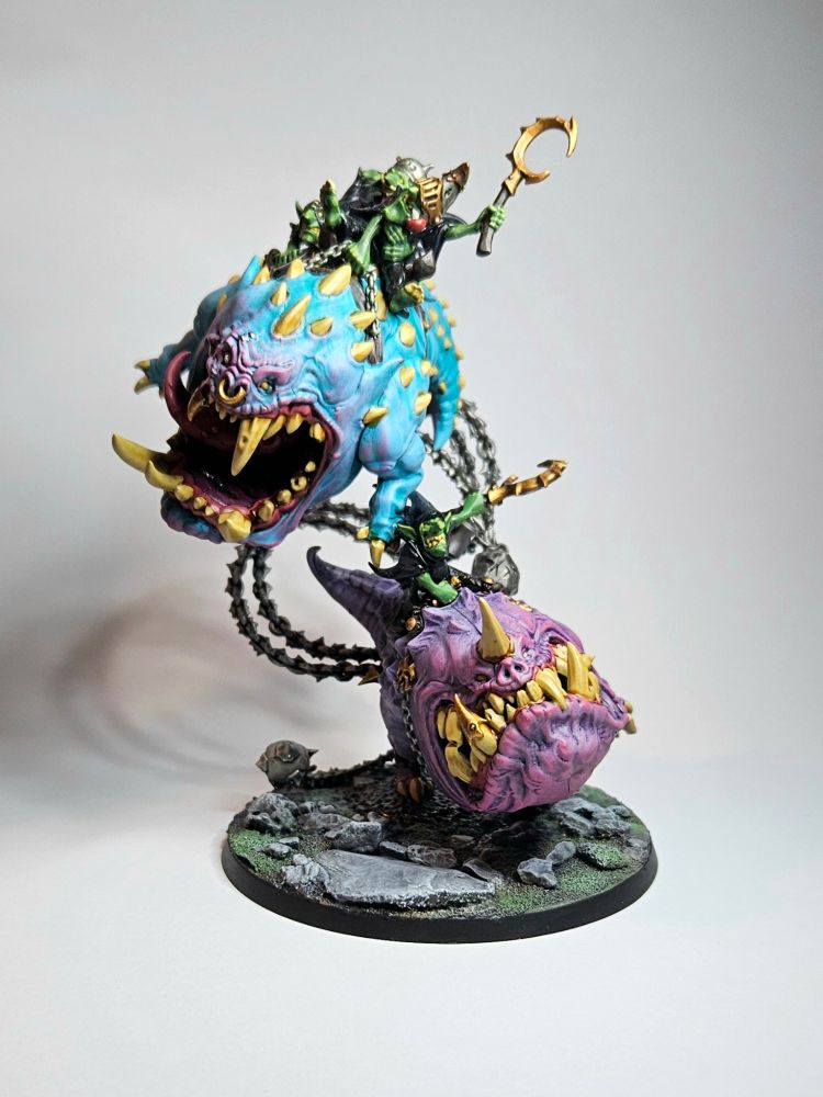 A pair of Warhammer Age of Sigmar Mangler Squigs, orb like toothy creatures. The lower one is purple with pink highlights and the upper one is blue with pink highlights. Riding them are various black robed goblins who are holding on for dear life!